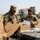 Two U.S. Army soldiers in MultiCam/OCP camouflage uniforms are inspecting the contents of a cardboard box and reviewing documents at what appears to be a forward operating base or logistics area. The male soldier on the left is opening the box while the female soldier on the right examines a white document or envelope. The setting appears to be an arid, dusty environment consistent with a Middle Eastern or desert deployment location.
