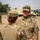 Two U.S. Army soldiers in Operational Camouflage Pattern (OCP) uniforms are engaged in a conversation at what appears to be a military installation or exercise area. The soldier facing away has the name tape 'NISBET' visible, while the soldier facing the camera bears the name tape 'KWON' and 'U.S. ARMY' tape. Military vehicles with camouflage paint are visible in the background.