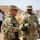 Two U.S. Army soldiers in MultiCam (OCP) uniforms and body armor are engaged in conversation at what appears to be a forward operating base or logistics site in an arid environment. Name tapes identify the soldiers as 'Luna' (left) and 'Saunders' (right), both wearing rank insignia consistent with senior non-commissioned officers. The background shows additional personnel, shipping containers, and stacked supplies suggesting a military logistics or humanitarian operation.