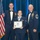 Airman Leadership School distinguished graduate Three United States Air Force personnel pose for a formal photograph at what appears to be an Airman Leadership School graduation ceremony. A female airman in the center holds a diploma from the United States Air Force Airman Leadership School, flanked by two senior enlisted members in dress uniforms with bow ties. The setting includes an American flag and Air Force flags against a blue curtain backdrop.