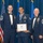 Airman Leadership School distinguished graduate Three United States Air Force personnel pose for a formal photo at what appears to be an Airman Leadership School graduation ceremony. The central figure holds a diploma framed in wood and wears a medal on a blue ribbon around his neck. The two flanking individuals wear dress blues with bow ties and display multiple service ribbons and rank insignia.