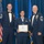 Airman Leadership School distinguished graduate Three United States Air Force personnel pose for a formal photograph during what appears to be a military graduation or awards ceremony. The central figure, a female airman, holds a diploma/certificate and wears a medal around her neck, flanked by two senior enlisted members in dress uniforms with bow ties. The setting includes an American flag and Air Force flags against a blue draped backdrop.