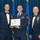 Airman Leadership School distinguished graduate Three United States Air Force personnel pose for a formal photograph at an Airman Leadership School diploma ceremony. The central figure holds a framed diploma from the United States Air Force Airman Leadership School, flanked by two senior enlisted members in mess dress uniforms. The setting includes American and Air Force flags against a blue curtain backdrop.