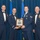 Airman Leadership School John L. Levitow awardee Four military personnel in U.S. Air Force dress uniforms pose for a formal award ceremony photograph. Two individuals in the center are jointly holding a wooden plaque featuring a portrait engraving, identified as the 'John L. Levitow Award.' The setting includes U.S. and Air Force flags against a blue draped backdrop, indicating a formal military banquet or ceremony.