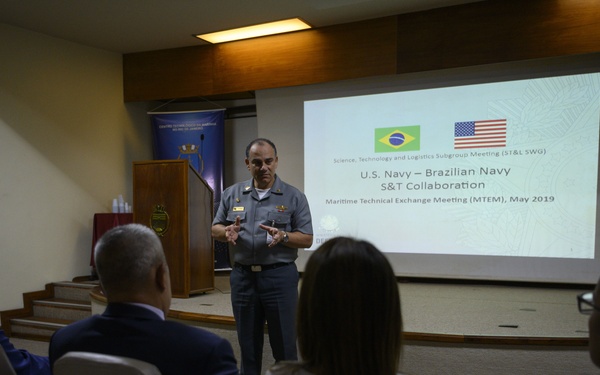 A military officer in uniform is presenting at a formal meeting between the U.S. Navy and Brazilian Navy, focused on Science and Technology collaboration. The presentation slide visible on the projector screen identifies this as a 'Maritime Technical Exchange Meeting (MTEM), May 2019.' Four attendees are seated in chairs facing the presenter in what appears to be a small auditorium or conference room.