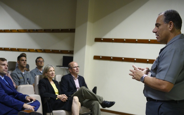 A military officer in a gray uniform is addressing a small group of civilians and uniformed personnel seated in an auditorium or briefing room. The setting appears to be a formal military or government facility, with the speaker gesturing with his hands as he presents. The audience is attentive and seated in theater-style chairs.