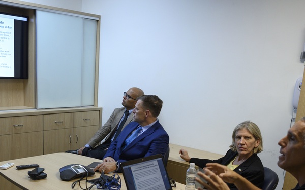 A small group of five professionals are gathered in a compact conference room for a presentation meeting. A projected slide on the wall TV displays text titled 'A (very) brief presentation about the collaboration between ONR and our group so far.' One participant appears to be speaking while others listen, with a laptop open on the conference table.