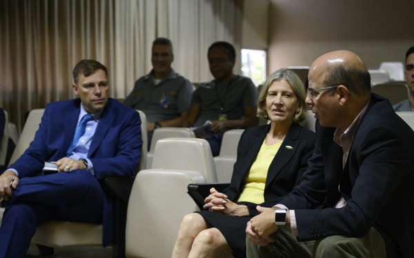 A formal meeting or briefing is taking place in what appears to be a conference or auditorium-style room. Several individuals in business attire are seated in rows of white chairs, with some engaged in conversation. The setting suggests an official or institutional gathering, with uniformed personnel visible in the background.