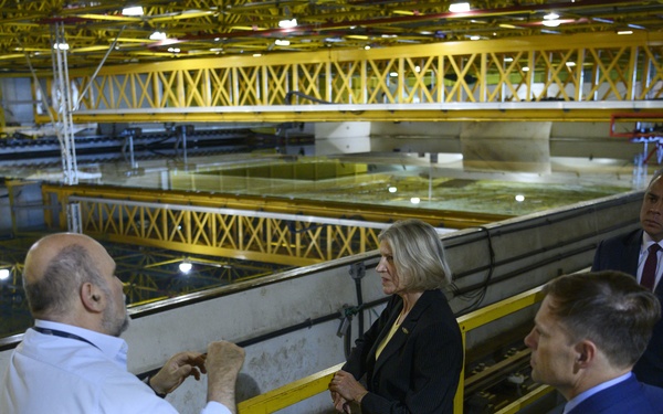 A group of five people are gathered on an elevated walkway or mezzanine level inside a large industrial facility, appearing to be on a guided tour or official visit. A man in a white shirt is gesturing and explaining something to the group, which includes formally dressed individuals. The facility below features large water tanks or basins with yellow overhead crane structures.