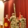 A U.S. Army military ceremony is taking place in an indoor auditorium or assembly hall with red curtains in the background. Soldiers in Army Combat Uniforms (ACU/OCP pattern) are participating in what appears to be a unit colors or guidon ceremony, likely a change of command or activation/deactivation event. The unit guidon visible is red with gold fringe and bears markings consistent with a Deployment and Distribution company.