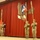 Four U.S. Army soldiers in OCP (Operational Camouflage Pattern) uniforms stand in a color guard formation on a stage with red curtains. They are holding military flags and guidons during what appears to be a formal military ceremony. A painted mural on the curtain backdrop depicts an eagle with a shield bearing the letter 'A', consistent with U.S. Army insignia.