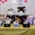 A display table covered with a yellow tablecloth holds multiple gift boxes and bags containing stuffed animals and baby items, arranged in front of a bulletin board. The black gift boxes are labeled 'with all my heart' and the boxes on the table appear to be sorted by weight categories. The setting appears to be a hospital or medical facility, likely related to a baby or infant donation/charity event.
