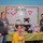 Five women are posing in front of a decorated bulletin board labeled 'L&amp;D 4DX' (Labor &amp; Delivery unit) with 'TELE TRAINED' written on it, suggesting a healthcare workplace celebration or recognition event. A table in front is decorated with a yellow tablecloth and displays gift boxes, stuffed animals, and other items. The setting appears to be a hospital or medical facility break room or common area.