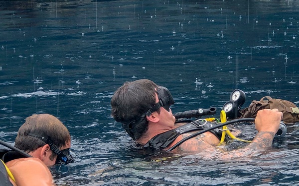 Two scuba divers are floating at the water surface, appearing to interact with each other or check equipment. Both are equipped with full scuba gear including tanks, BCDs, regulators, and masks pushed up on their foreheads. The water appears choppy with visible rain droplets hitting the surface, suggesting outdoor conditions during light rainfall.