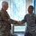 Two U.S. military personnel are shaking hands in what appears to be an indoor facility with large windows and vertical blinds in the background. The individual on the left wears an OCP (Operational Camouflage Pattern) uniform, while the individual on the right wears an ABU (Airman Battle Uniform) with 'U.S. AIR FORCE' tape and a Staff Sergeant rank insignia visible. The interaction appears to be a formal recognition or greeting ceremony.