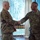 Two military personnel in OCP (Operational Camouflage Pattern) uniforms are shaking hands in what appears to be an indoor facility with large windows and vertical blinds in the background. The individual on the right has a visible name tape and 'U.S. AIR FORCE' tape, along with what appears to be a senior NCO rank insignia. The handshake suggests a formal recognition, greeting, or ceremonial exchange.