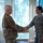 Two military personnel are shaking hands in what appears to be an indoor ceremony or formal meeting. The individual on the left wears an Army Combat Uniform (OCP/Multicam) with a unit patch and U.S. flag, while the individual on the right wears a U.S. Air Force Airman Battle Uniform (ABU) with visible rank insignia and a name tape. The setting appears to be an office or meeting room with vertical blinds and windows overlooking trees.
