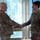 Two military personnel in OCP (Operational Camouflage Pattern) uniforms are shaking hands in what appears to be an indoor military facility. An older male service member on the left is shaking hands with a younger female service member on the right, who is smiling. The setting and body language suggest a formal recognition, promotion, or award ceremony.