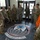 A group of approximately nine military personnel in various uniforms are gathered in the lobby of what appears to be the 212th Rescue Squadron facility. One individual on the right appears to be briefing or presenting to the group, while a mannequin dressed in rescue/flight gear with an orange survival suit is visible in the foreground. The floor features the 212th Rescue Squadron emblem with the motto 'THAT OTHERS MAY LIVE'.