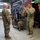 Two military personnel in MultiCam/OCP camouflage uniforms are engaged in a briefing or equipment review inside what appears to be a gear storage facility. The soldier on the right is gesturing with both hands, apparently explaining or demonstrating something to the senior soldier on the left. The background contains a wire shelving rack loaded with tactical gear, bags, and various outdoor/special operations equipment.