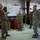 Military personnel are gathered inside what appears to be a maintenance hangar or equipment bay for a briefing or status update. A senior soldier on the left is gesturing toward a whiteboard displaying a tracking chart or status board, while others listen attentively. The group includes personnel in both OCP (Operational Camouflage Pattern) and ABU/Airman Battle Uniform patterns, suggesting a joint or multi-unit gathering.