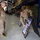 Two military personnel in OCP (Operational Camouflage Pattern) uniforms are inside a warehouse or hangar facility. One soldier is pointing to an A-frame display board containing informational charts, photos, and text, while the other observes. The background contains military equipment including packed parachutes or rucksacks on blue metal shelving racks.