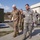 A group of military personnel in uniform are walking together outdoors at what appears to be a military installation or base. The foreground shows two senior officers — one in U.S. Army OCP (Operational Camouflage Pattern) and one in U.S. Air Force ABU (Airman Battle Uniform) — engaged in conversation during what appears to be an official tour or inspection. Several additional uniformed personnel follow behind them.
