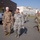 A group of military personnel in various camouflage uniforms are walking across a paved area at an Alaska Air National Guard facility. The foreground shows two senior personnel engaged in conversation, with additional uniformed individuals following behind. The building in the background is clearly marked 'Alaska Air National Guard' with a secondary sign reading 'Home of the 176th Wing'.