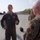 Military personnel are gathered on an airfield tarmac engaged in conversation. A pilot in an olive flight suit with Air Force patches is speaking to a senior officer in OCP (Operational Camouflage Pattern) uniform. Military helicopters, likely HH-60 Pave Hawks, are visible in the background.