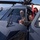 Two military personnel are seated in the cockpit of a gray military helicopter, with one individual appearing to explain or point out features to the other. The helicopter is parked on what appears to be a flight line or tarmac near a hangar facility. A third person is partially visible in the background to the right.