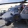 A gray military helicopter, marked with the number '7068' on its nose, is parked on a tarmac apron outside a hangar facility. Two individuals in military attire are engaged in what appears to be a briefing or inspection near the open cockpit doors. The helicopter appears to be a Sikorsky HH-60 Pave Hawk or similar Black Hawk variant based on its configuration.