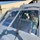 Two military personnel are seated in the cockpit of a military helicopter, likely a UH-60 Black Hawk, on a tarmac or airfield. The person in the right seat is giving a thumbs-up gesture toward the camera while wearing a flight helmet with a microphone. A large military hangar building is visible in the background.