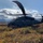 A military helicopter, consistent with a Sikorsky HH-60 Pave Hawk or UH-60 Black Hawk variant, is landed on open tundra terrain with autumn-colored low shrubs. The aircraft is painted in a dark olive/gray military finish and appears to be equipped with an aerial refueling probe extending from the nose. The setting appears to be a remote Alaskan or subarctic wilderness, with mountain ranges visible in the background under a partly cloudy sky.
