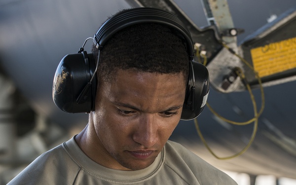 A military or aviation ground crew member wearing hearing protection (ear defenders) reviews documents or forms while standing near an aircraft. He is dressed in a tan/sage military-style physical training shirt and holds what appears to be a stack of maintenance or inspection paperwork. The setting appears to be an outdoor flight line or tarmac area.