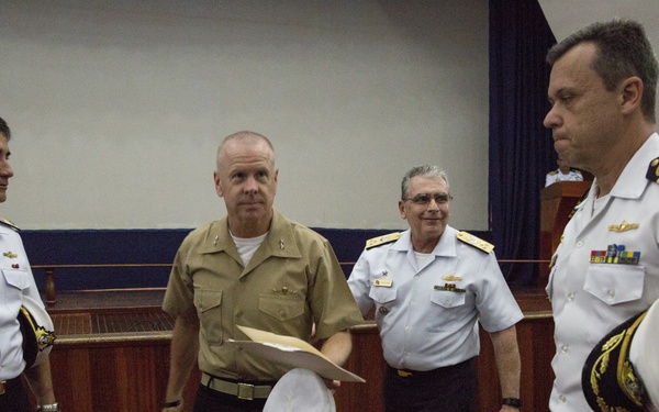 A group of military naval officers are gathered in what appears to be a formal meeting room or auditorium. The officers are wearing a mix of uniforms including tan/khaki and white naval dress uniforms with rank insignia and service ribbons. Several officers are holding white naval dress caps, suggesting a formal ceremony or meeting has recently concluded or is about to begin.