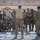 A group of military personnel in mixed camouflage uniforms are gathered outdoors in what appears to be a briefing or training session. A central figure with his back to the camera appears to be addressing the assembled group. Multiple water bottle packs are placed on the ground, suggesting a hot-weather outdoor training environment.