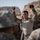 A military instructor in MultiCam/OCP camouflage uniform is conducting a training demonstration outdoors in an arid, rocky terrain. He is holding up two circular objects in both raised hands, apparently instructing a group of soldiers gathered around him. The setting appears to be a field training exercise in a desert or semi-arid environment.