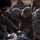 Several military personnel in camouflage uniforms are gathered around what appears to be a litter or stretcher, engaged in what looks like a medical training or casualty care exercise. The central figure wears a multicam uniform with a cap bearing a military insignia patch and a blood type patch (A+) on the chest. The setting appears to be an outdoor arid environment, suggesting a field training exercise.