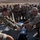 A group of military personnel in camouflage uniforms are gathered outdoors in what appears to be a training or demonstration exercise. Two soldiers in the foreground are handling equipment cases and gear on a surface, while a large group of uniformed observers watches from behind. The setting appears to be a military base or training facility in an arid environment.