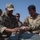 Two military personnel in camouflage uniforms are collaborating over what appears to be equipment or a document on a surface, in an arid outdoor environment. One soldier wears a patch consistent with Jordanian Armed Forces insignia, while the other displays a U.S. flag patch, suggesting a joint military training or coordination exercise. Additional personnel are visible in the background observing the interaction.