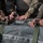 Multiple individuals in military camouflage uniforms are working together to secure or adjust straps and D-ring hardware on a large gray tactical bag or equipment bundle. The close-up shot focuses on their hands manipulating olive-drab webbing straps with metal D-rings. The activity appears to be rigging or preparing equipment, possibly for aerial delivery or transport.