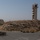 A group of military personnel in uniform are assembled on a paved road or tarmac area, moving toward a convoy of military tactical vehicles. The scene takes place in an arid, desert-like environment featuring a tall concrete observation or control tower under construction on a rocky hill. The setting appears to be a military base or training facility in a dry, mountainous region.