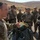 A U.S. Army soldier, identifiable by the American flag patch on his left shoulder, is conducting a briefing or instruction session for a group of foreign military personnel. He is gesturing toward what appears to be a packed parachute or military equipment on the ground. The setting appears to be an arid, outdoor military training environment.