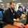 A bearded man wearing a dark jacket with a FEMA logo is seated at a workstation in what appears to be an emergency operations center, looking toward the camera. Multiple personnel are visible working at computer stations in the background. The environment is a large, open office-style operations room with fluorescent lighting and multiple monitors.