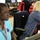 The image shows multiple people working at computer workstations in what appears to be an operations center or command room. The foreground features a woman wearing a teal polo shirt with a badge/patch and a 'Cowboys' lanyard, focused on a monitor. Several other workers are visible in the background, also engaged at their workstations.