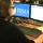 A person with shoulder-length hair and glasses is seated at a desk working at a dual-monitor Dell computer workstation displaying a FEMA (Federal Emergency Management Agency) desktop background. The left monitor shows the FEMA logo and seal with a notice about document management, while the right monitor displays an active web browser or application. The setting appears to be an emergency operations or coordination center.