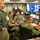 Military and emergency management personnel are gathered in what appears to be a FEMA National Response Coordination Center (NRCC), as indicated by visible signage. Multiple service members in various uniforms — including Navy digital camouflage, Army ACU, and Air Force — are working at computer workstations monitoring what appear to be weather or disaster-related data. The large video wall in the background displays maps and satellite imagery consistent with hurricane or major storm tracking operations.
