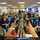 The image shows a large emergency operations center or command center with numerous workstations equipped with multiple monitors. Staff members are actively engaged in conversations and working at computer terminals arranged in long rows. The facility features large display screens on the walls and appears to be in a state of active operation.