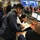 The image shows personnel working inside a FEMA (Federal Emergency Management Agency) operations center. Multiple staff members are engaged in various tasks including reviewing documents, monitoring computer workstations, and coordinating activities. Large display screens showing data and weather maps are visible in the background.