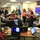 The image shows an emergency operations center, likely a FEMA National Response Coordination Center (NRCC), with multiple personnel actively engaged in a briefing or coordination meeting. Staff members wearing various agency vests and badges, including FEMA and NRCS identifiers, are gathered around workstations with laptops and documents. A large display board showing time zones and status information is visible in the background.